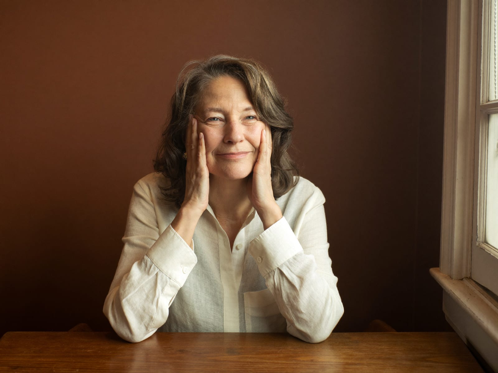 A woman in her fifties sits at a wooden table beside a window in soft warm light, both hands resting against her cheeks with the fingers lifting them into a closed-mouth smile, wearing an ivory linen shirt against a plain espresso-brown wall, calm and unposed, natural texture visible in her skin.