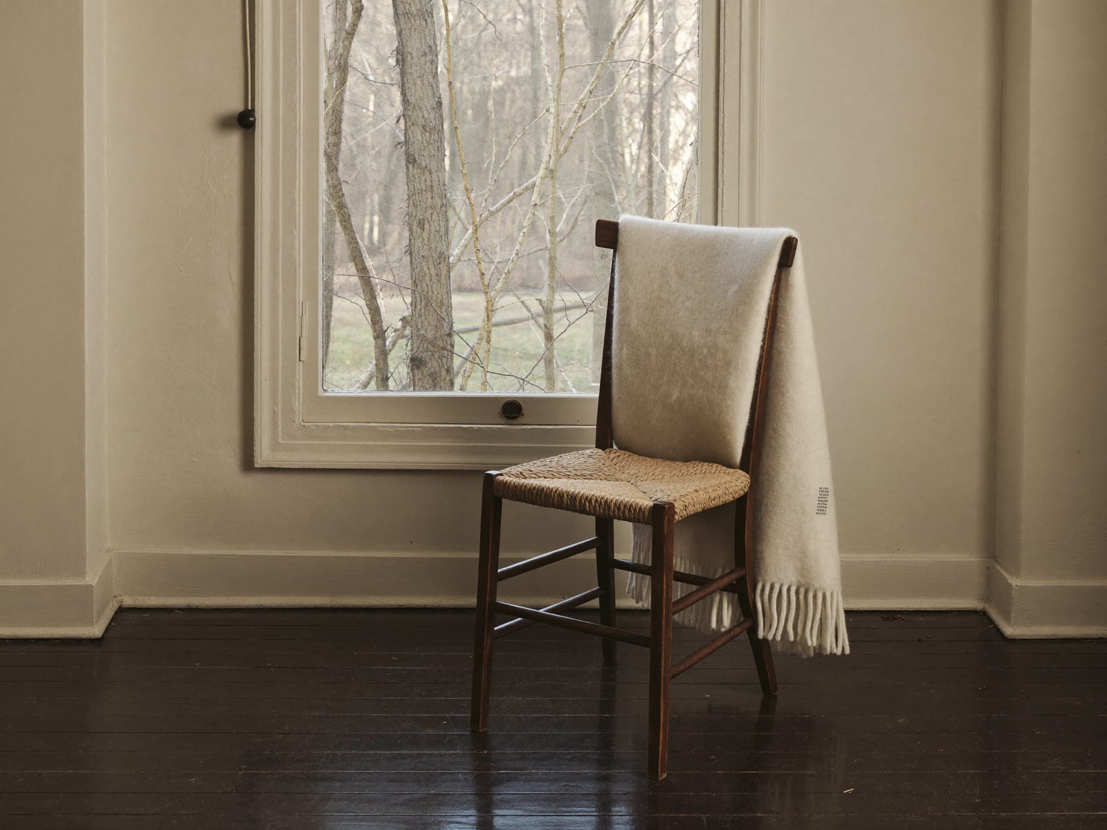 An empty dark wooden chair with a rush seat standing beside a window on a grey autumn morning, a cream wool blanket draped over its back and hanging to the floor, bare trees showing through the glass, ivory plaster walls and dark espresso floorboards, nobody in the room, warm ivory and espresso palette, 4:3.