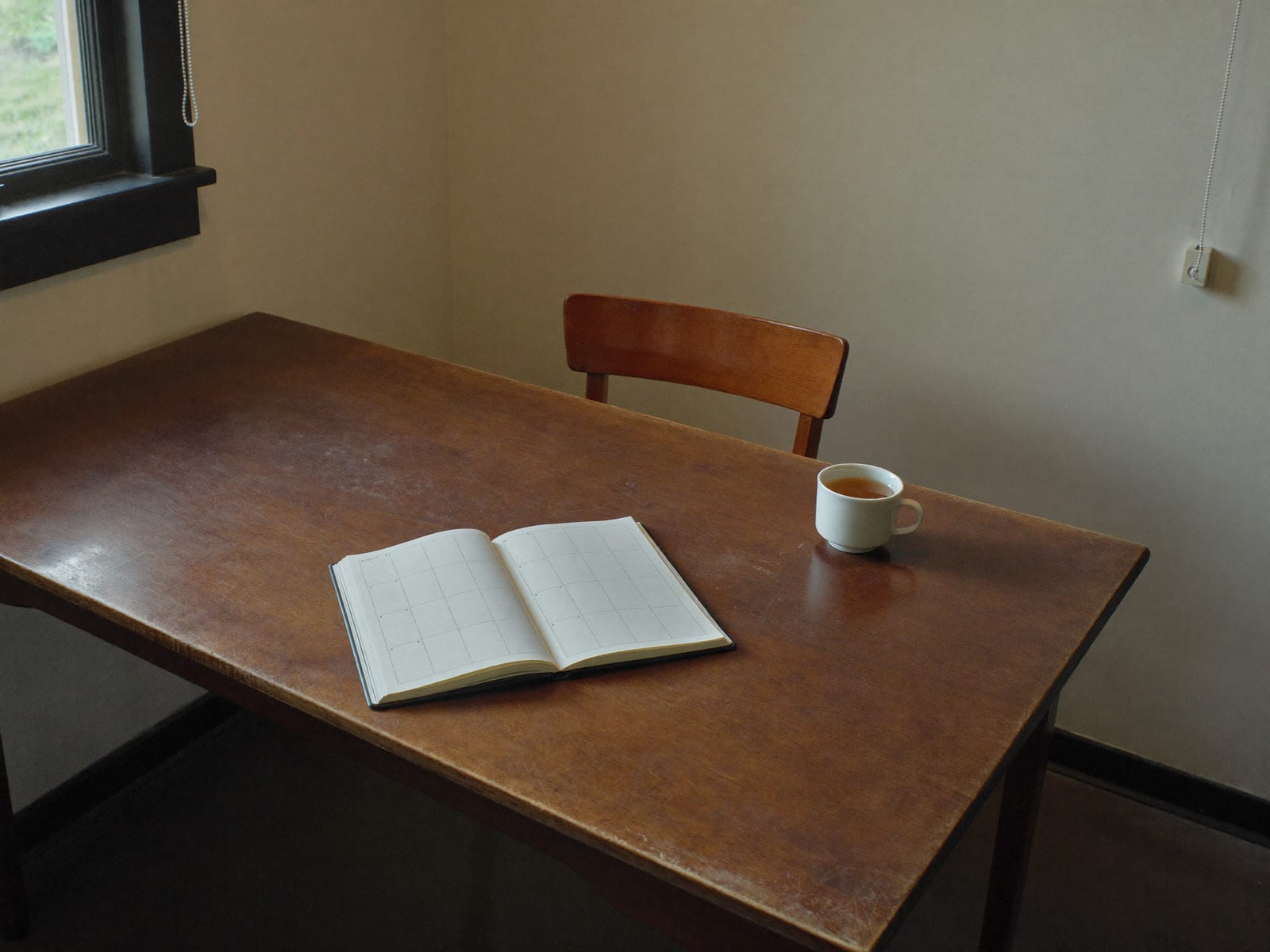 A worn wooden table in the corner of a plain room, an open paper diary lying flat on it beside a white cup of tea, one chair pushed back and nobody in the room, daylight coming in from a window at the left, warm ivory and espresso palette, soft natural light, 4:3, no text.