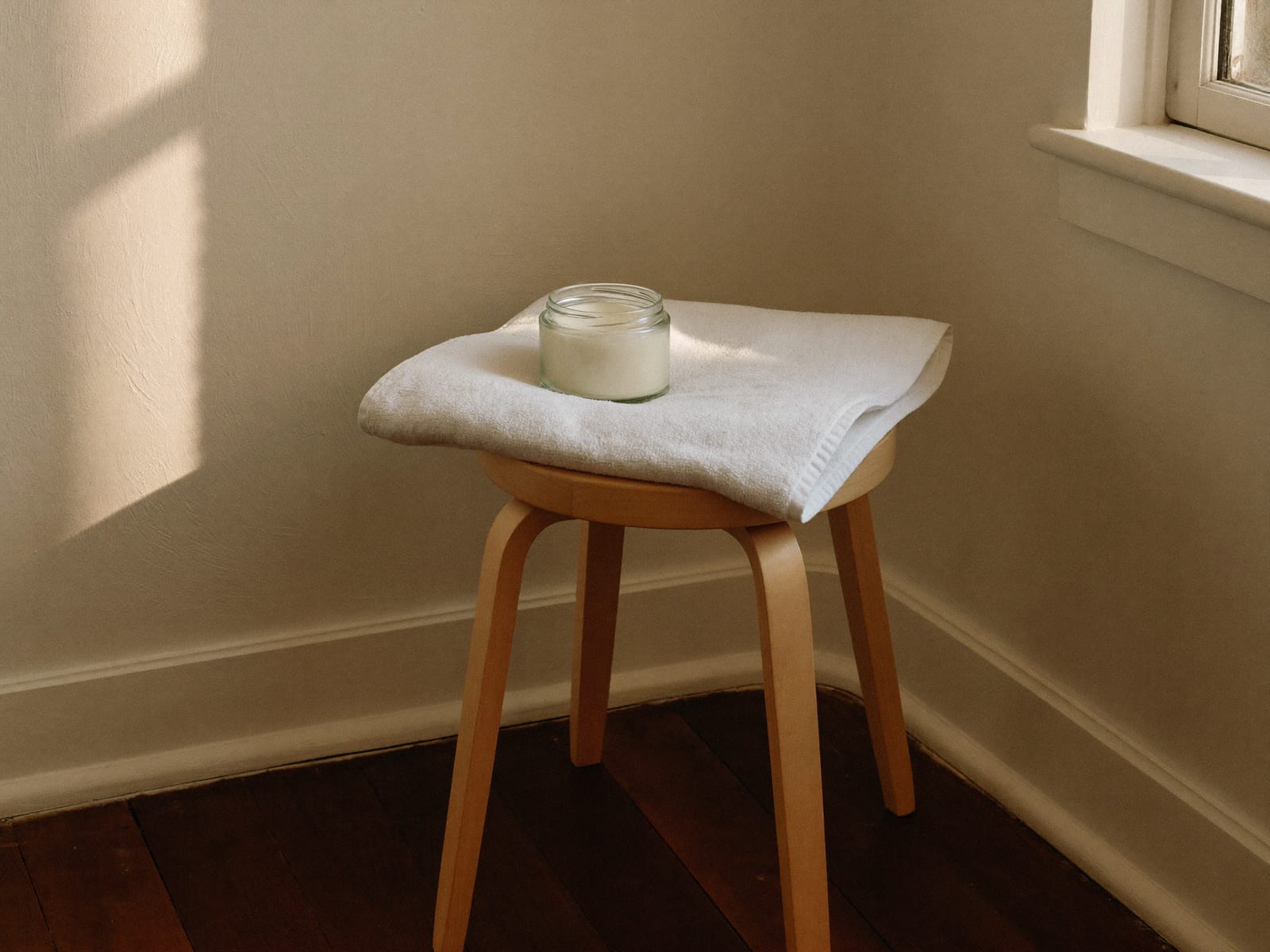 A white cotton towel draped over a pale birch stool with a plain glass jar of white ointment sitting on it, morning light from a window falling in a soft shaft across an ivory plaster wall, dark espresso floorboards below, nobody in the room, warm ivory and espresso palette, 4:3, no text.